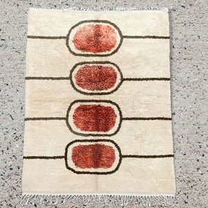May include: A rectangular area rug with a cream-coloured base featuring four horizontally aligned, dark brown outlined ovals. Each oval contains a textured, burnt orange centre. The rug has a knotted fringe along the top and bottom edges.
