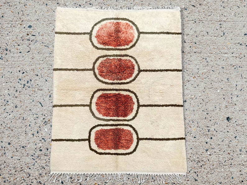 May include: A rectangular area rug with a cream-coloured base featuring four horizontally aligned, dark brown outlined ovals. Each oval contains a textured, burnt orange centre. The rug has a knotted fringe along the top and bottom edges.