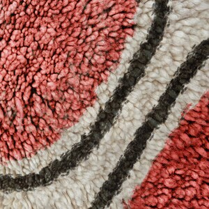 May include: A close-up view of a textured rug featuring thick, looped pile in shades of coral pink and cream, separated by two distinct, curved dark charcoal grey lines that create a flowing, organic pattern across the surface.