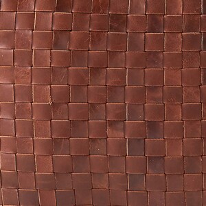 May include: A close-up view of a brown leather surface featuring a woven pattern. The material consists of thin, flat strips of leather interlaced in a grid formation, creating a uniform, square-tiled texture across the entire visible area.