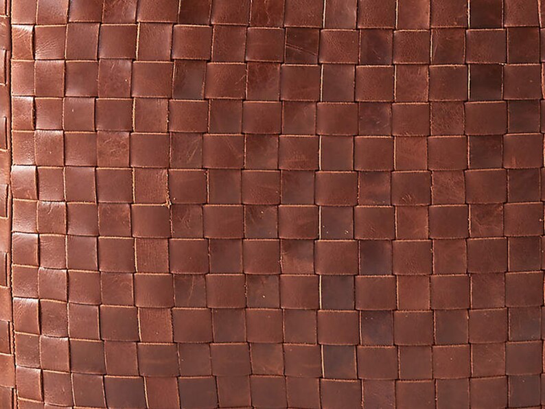 May include: A close-up view of a brown leather surface featuring a woven pattern. The material consists of thin, flat strips of leather interlaced in a grid formation, creating a uniform, square-tiled texture across the entire visible area.