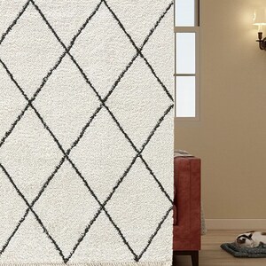 May include: A plush, off-white area rug featuring a repeating black diamond grid pattern. The rug has a thick, textured pile and a fringed edge at the bottom, creating a soft surface with a high-contrast geometric design.
