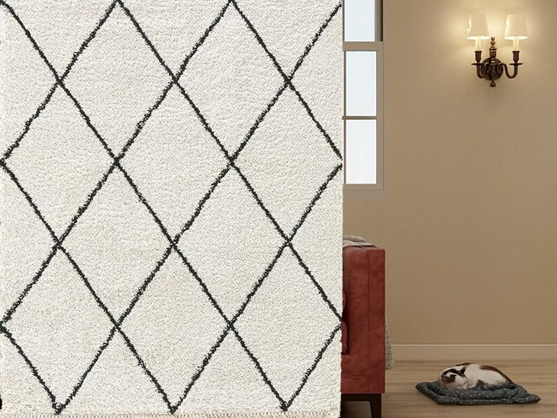 May include: A plush, off-white area rug featuring a repeating black diamond grid pattern. The rug has a thick, textured pile and a fringed edge at the bottom, creating a soft surface with a high-contrast geometric design.