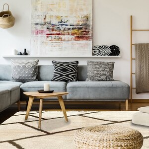 May include: A cream-colored area rug featuring a dark brown geometric grid pattern. The rug has a thick, textured pile and is positioned in a living room setting with a gray sectional sofa, a wooden coffee table, and various decor items.