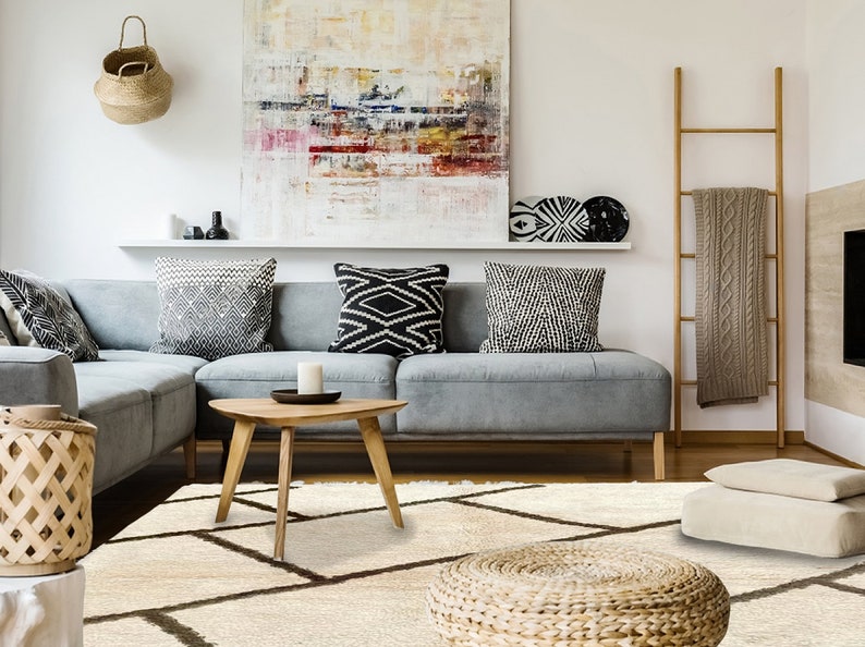 May include: A cream-colored area rug featuring a dark brown geometric grid pattern. The rug has a thick, textured pile and is positioned in a living room setting with a gray sectional sofa, a wooden coffee table, and various decor items.