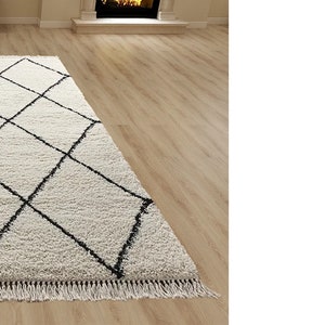 May include: A rectangular area rug with a thick, cream-colored shaggy pile featuring a repeating black diamond grid pattern. The rug is finished with short, cream-colored fringe along the edge, creating a textured surface appearance.
