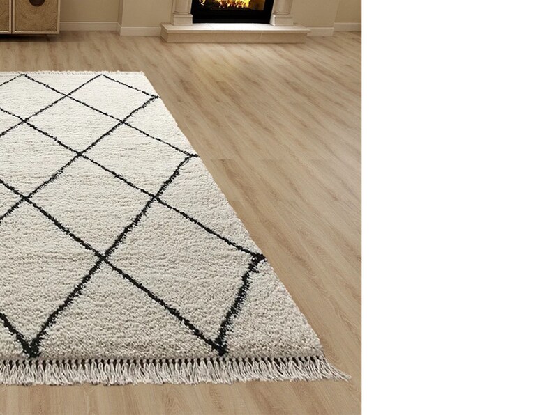 May include: A rectangular area rug with a thick, cream-colored shaggy pile featuring a repeating black diamond grid pattern. The rug is finished with short, cream-colored fringe along the edge, creating a textured surface appearance.