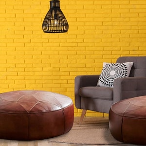 Op de afbeelding: Twee ronde, bruine lederen poefs met gestikte geometrische panelen, geplaatst voor een grijze gestoffeerde fauteuil met een wit kussen met een zwart cirkelvormig patroon, tegen een felgele bakstenen muur.