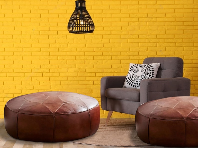 Op de afbeelding: Twee ronde, bruine lederen poefs met gestikte geometrische panelen, geplaatst voor een grijze gestoffeerde fauteuil met een wit kussen met een zwart cirkelvormig patroon, tegen een felgele bakstenen muur.