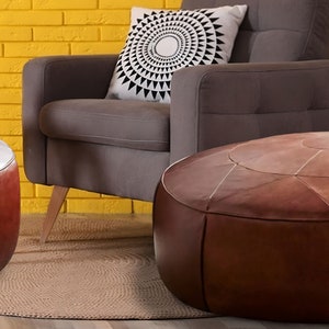 Op de afbeelding: Twee ronde, bruine lederen poefs met gestikte paneeldetails, geplaatst voor een grijze gestoffeerde fauteuil met een wit kussen met een zwart geometrisch zonnestraalpatroon tegen een gele bakstenen muur.