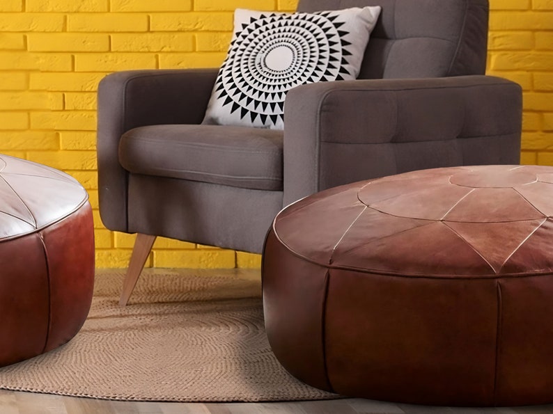 Op de afbeelding: Twee ronde, bruine lederen poefs met gestikte paneeldetails, geplaatst voor een grijze gestoffeerde fauteuil met een wit kussen met een zwart geometrisch zonnestraalpatroon tegen een gele bakstenen muur.