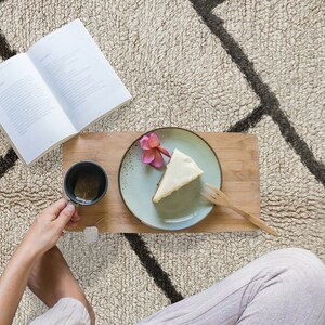 May include: A rectangular bamboo serving tray holds a light green ceramic plate with a slice of cheesecake topped with a pink flower, a small black mug containing tea with a tea bag, and a wooden fork. An open book lies nearby.