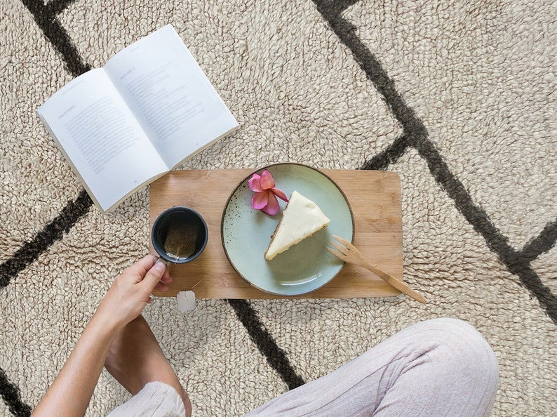 May include: A rectangular bamboo serving tray holds a light green ceramic plate with a slice of cheesecake topped with a pink flower, a small black mug containing tea with a tea bag, and a wooden fork. An open book lies nearby.