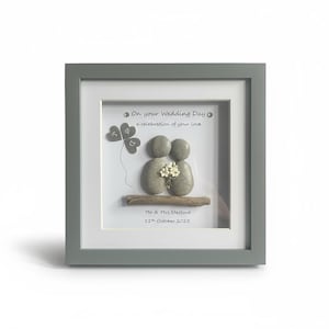 May include: A square grey framed wall art piece featuring pebble art of two figures sitting on a piece of driftwood. The figures hold white flowers. Text reads: On your wedding day, a celebration of your love, Mr & Mrs Stafford, 11th October 2025.