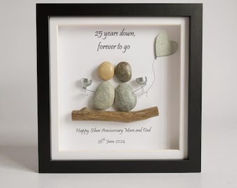 May include: A framed pebble art piece featuring two figures made of stones sitting on a wooden branch. The figures hold small silver cups, and a heart-shaped stone balloon floats above. Text reads: 25 years down, forever to go, Happy Silver Anniversary Mam and Dad, 26th June 2024.