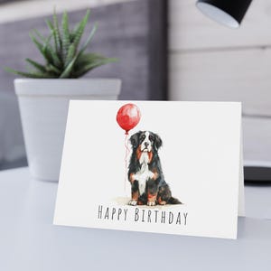 May include: A white greeting card featuring a watercolor illustration of a Bernese Mountain Dog sitting and holding a red balloon by its string. The text HAPPY BIRTHDAY is printed in black capital letters at the bottom of the card front.
