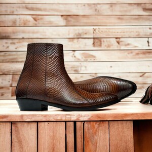 May include: Brown leather ankle boots with a reptile-like texture. The boots have a pointed toe and a black heel. The boots are displayed on a wooden surface. The boots are approximately 15 cm high.