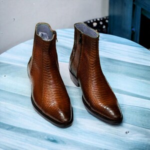 May include: A pair of brown leather ankle boots with a snakeskin texture. The boots have a side zip and a pointed toe. The boots are on a light blue surface.