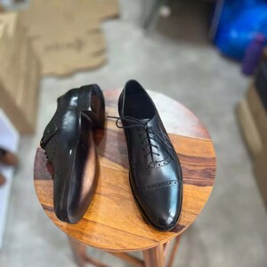 Pu&ograve; includere: Un paio di scarpe stringate in pelle nera con motivo brogue. Le scarpe sono su una superficie di legno.
