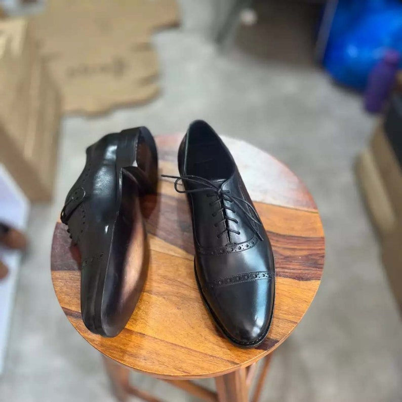Pu&ograve; includere: Un paio di scarpe stringate in pelle nera con motivo brogue. Le scarpe sono su una superficie di legno.