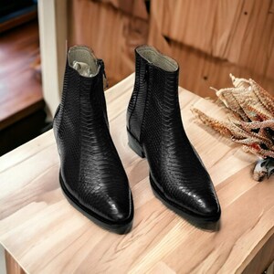 Könnte beinhalten: Ein Paar schwarze Leder-Ankle-Boots mit einem strukturierten, schlangenartigen Muster. Die Stiefel haben eine spitze Zehenpartie und einen Reißverschluss an der Seite. Sie werden auf einer hellen Holzoberfläche präsentiert.