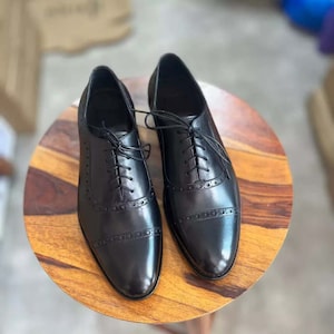Pu&ograve; includere: Un paio di scarpe stringate in pelle nera con motivo brogue. Le scarpe hanno punta a punta e sono appoggiate su una superficie in legno.