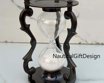 Può includere: Una clessidra in stile antico con finitura bronzo scuro. La lampadina di vetro contiene sabbia bianca, tenuta all'interno di una cornice metallica decorativa. La clessidra ha una parte superiore e inferiore circolari, con supporti curvi ornati.