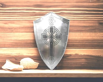 May include: A silver-colored shield with a Celtic cross design. The shield is in front of a wooden background. Two small, light-colored objects are at the bottom left of the shield.
