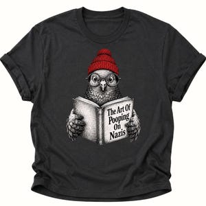Könnte beinhalten: Ein schwarzes T-Shirt mit einer grafischen Illustration eines Vogels, der eine rote Strickmütze und eine runde Brille trägt. Der Vogel hält ein offenes Buch, auf dessen Einband der Text The Art Of Pooping On Nazis in Schwarz steht.