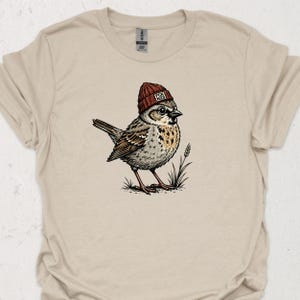 Op de afbeelding: Een zandkleurig t-shirt met een grafische illustratie van een kleine bruine vogel die een donkerrode gebreide muts draagt. De muts heeft een kleine witte patch met de tekst FCK NZS. De vogel staat bij een kleine grasspriet of graanhalm.
