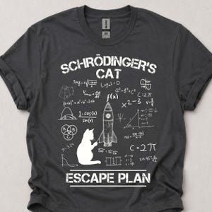 Könnte beinhalten: Dunkelgraues T-Shirt mit weißem Aufdruck. Das Design zeigt den Text "SCHRÖDINGER'S CAT ESCAPE PLAN", mathematische Formeln, eine Rakete und eine Katzen-Silhouette. Das T-Shirt ist aus weichem Stoff.