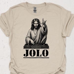 Könnte beinhalten: Ein beiges T-Shirt mit kurzen Ärmeln und einem schwarzen Grafikdruck einer bärtigen Figur, die Kopfhörer trägt und ein DJ-Mischpult bedient. Die Figur macht ein Handzeichen und der Text JOLO ist in großen schwarzen Buchstaben unter dem Pult gedruckt.