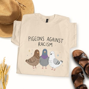 Könnte beinhalten: Beiges T-Shirt mit dem Text "Pigeons Against Racism" und einer Grafik von drei Tauben in Braun, Grau und Weiß. Im Hintergrund sind ein Strohhut und Sandalen zu sehen.