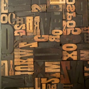 May include: A collection of vintage wooden letterpress printing blocks. The blocks are a mix of capital letters, numbers, and symbols in various sizes and fonts. The wood has a warm, natural tone, contrasting with the dark background.