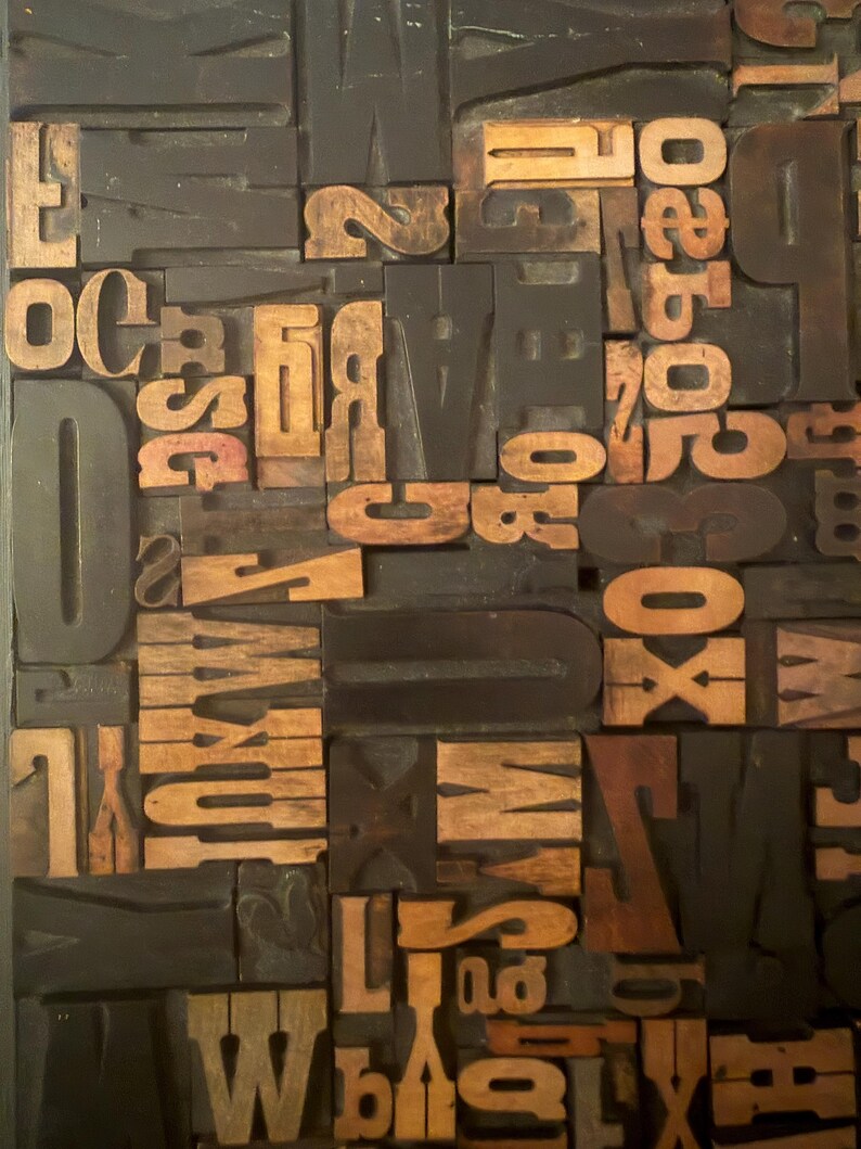 May include: A collection of vintage wooden letterpress printing blocks. The blocks are a mix of capital letters, numbers, and symbols in various sizes and fonts. The wood has a warm, natural tone, contrasting with the dark background.