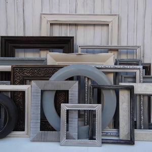 May include: A collection of various picture frames in different shapes, sizes, and colors. The frames include black, white, gray, and brown, with ornate and simple designs. Some frames are round, square, and rectangular.