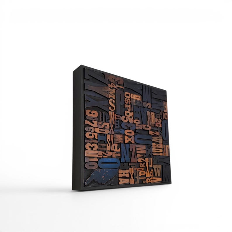 May include: A framed artwork featuring a collage of vintage wooden letterpress printing blocks. The blocks are in various sizes and fonts, with a mix of black, blue, and brown tones. The frame is black.