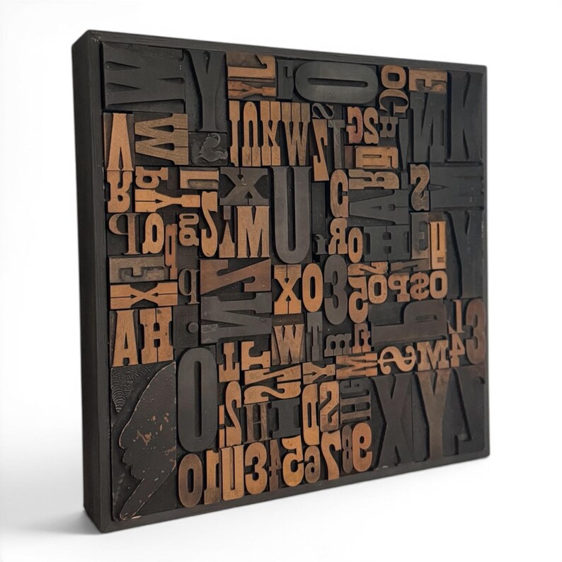 May include: A square, wooden block art piece featuring an assortment of vintage letterpress printing blocks. The blocks are a mix of dark brown and natural wood tones, arranged in a seemingly random pattern within a black frame.