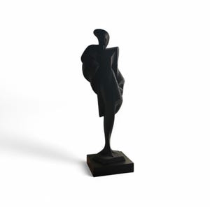 May include: A black abstract sculpture of a figure in a draped garment, standing on a square base. The sculpture has a minimalist design and is set against a white background. The figure's form is elongated and stylized.