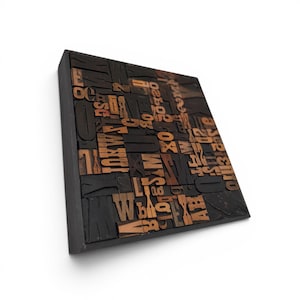 May include: A square, black wooden block with a collection of vintage wooden letterpress printing blocks. The letters and numbers are in various sizes and fonts, with a warm brown color. The block is mounted on a white background.