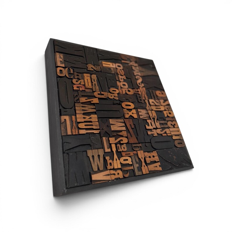 May include: A square, black wooden block with a collection of vintage wooden letterpress printing blocks. The letters and numbers are in various sizes and fonts, with a warm brown color. The block is mounted on a white background.