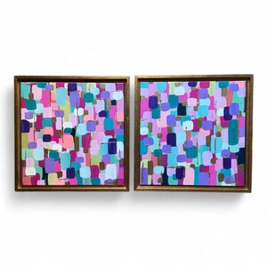 Peut inclure: Deux peintures abstraites carr&eacute;es avec des cadres dor&eacute;s. Chaque &oelig;uvre pr&eacute;sente un arrangement color&eacute; de formes rectangulaires dans des tons de rose, de violet, de bleu et d'or. Les peintures sont expos&eacute;es sur un fond blanc.