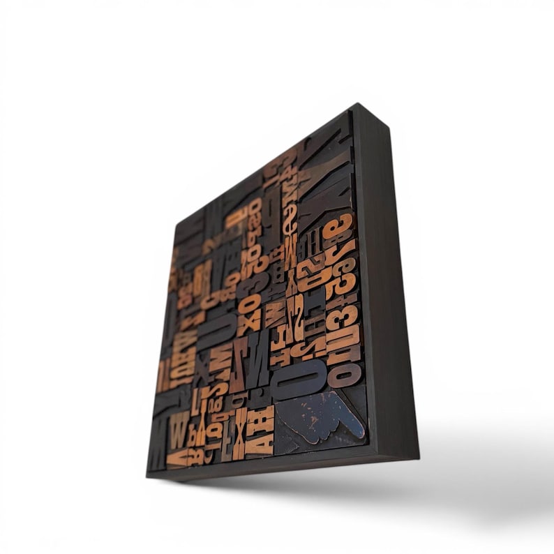 May include: A square, dark wooden wall art piece featuring a collage of vintage letterpress printing blocks. The blocks are various sizes and fonts, with a mix of black, brown, and tan colors. A pointing hand is visible.