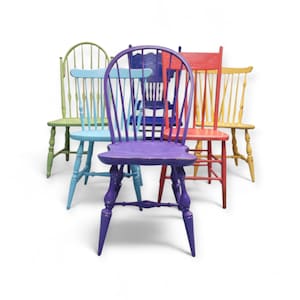 May include: A collection of five brightly painted wooden chairs. Each chair is a different color: green, blue, purple, red, and yellow. The chairs have spindle backs and turned legs, and are arranged in a semi-circle.