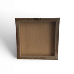 May include: Back view of a square wooden framed art piece. The frame is a dark color with a lighter, distressed edge. The back of the artwork is a light brown wood, with a metal hanger attached at the top.