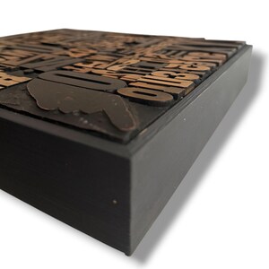 May include: A vintage letterpress block with copper-colored letters and symbols set in a black wooden frame. The block includes a pointing hand and the number "0". The letters are arranged in a seemingly random pattern.