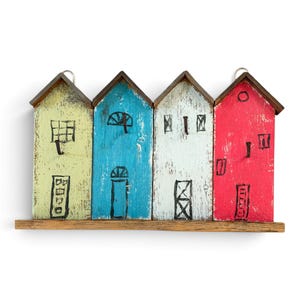 May include: A wall-mounted wooden key holder featuring four house-shaped panels in light green, bright blue, white, and red. Each panel has a dark brown roof, a hand-drawn black ink illustration of doors and windows, and a small metal hook.