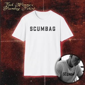 May include: A white short-sleeved t-shirt featuring the word SCUMBAG printed in a black, distressed, block-letter font across the chest. The text Josh Homme's Scumbag Tshirt appears in the top left corner of the image.
