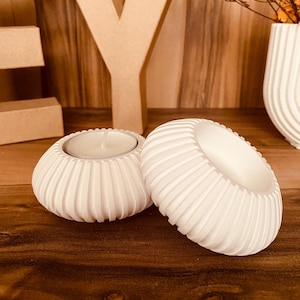 Könnte beinhalten: Zwei weiße, geriffelte Kerzenhalter aus Keramik. Einer enthält eine brennende Teelichtkerze. Die Kerzenhalter stehen auf einer Holzoberfläche, im Hintergrund eine dekorative Vase und Holzbuchstaben. Ein dekoratives Wohnaccessoire.