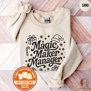 May include: A sand-colored sweatshirt with the words "Magic Maker Manager" in a black, whimsical font. The design includes snowflakes, gift boxes, and a Santa hat. The sweatshirt has a crew neck and long sleeves.